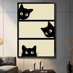 Black framed canvas print of three curious black cats peeking over edges of three beige horizontal panels in minimalist cartoon style, displayed on grey wall above console table with candles

