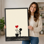 Smiling woman holding a black framed Banksy-style minimalist black figure touching a red heart plant art print on a cream background against a neutral indoor background with potted plants