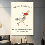 Gallery-wrapped stretched canvas of a chubby cartoon seagull holding a french fry with Happy Birthday and may you be as happy as a seagull with a french fry quote, funny illustrated bird print on off-white background, hung above a dark sideboard with candlesticks against a gray paneled wall