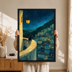 Close-up of a black-framed art print showing a black cat on an ornate wrought-iron balcony railing with a glowing street lantern and starry hillside village in the background
