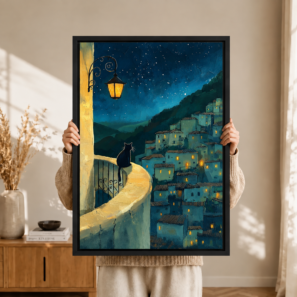 Close-up of a black-framed art print showing a black cat on an ornate wrought-iron balcony railing with a glowing street lantern and starry hillside village in the background
