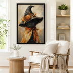 Framed gothic-whimsical wall art of a black cat wearing a tall witch hat decorated with fall pumpkins and maple leaves, styled beside a linen armchair in a warm Scandinavian reading nook
