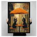 Hand-held framed print of two golden-eyed black cats at an autumn street café, orange umbrella and warm amber leaf-scattered alley background