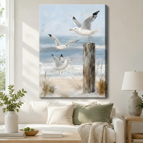 Stretched canvas coastal seagull wall art depicting three gulls soaring above a driftwood post on a sandy beach with soft blue ocean tones, hung above a white sofa in a modern coastal living room