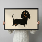 Person holding a black framed art print of a whimsical dachshund illustration with orange accents on a warm cream background, showing print size and quality