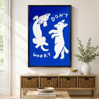Black framed bold blue "Don't Worry" dancing dogs wall art with white line illustration mounted above a light wood wicker basket console with ceramic bowls, stacked books, and a tall white vase in a warm minimal entryway
