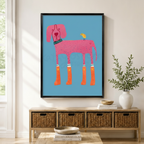 Funky pink dog wearing orange star rain boots with yellow bird on back black framed canvas art in sky blue above a rattan sideboard with wicker baskets, stacked books, and a white ceramic vase in a Scandinavian living room
