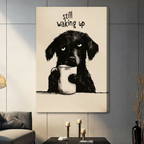 Humorous pencil illustration fluffy black dog clutching white coffee cup with tired eyes "Still Waking Up" gallery wrap canvas art on warm beige background on a dark gray paneled wall above a charcoal sofa with candle holders and a black vase