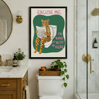 Humorous leopard bathroom wall art in a black frame with pink and green "Excuse Me I'm Kind of Busy In Here" text, hung above a toilet in a luxury bathroom with marble countertops, gold fixtures, and trailing ivy