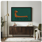Black framed cartoon long dachshund "It's Going to Be a Long Day" horizontal wall art in teal green above a dark walnut sideboard with stacked books and a succulent, beside a brass floor lamp and olive green armchair with a knit throw blanket in a modern living room