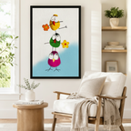 Black framed funny stacked birds with flowers canvas art showing three wide-eyed round cartoon birds — yellow on top, green in the middle, magenta at the bottom — stacked and balancing while holding orange and yellow daisies on a white and sky blue background, displayed beside a white linen armchair in a cozy reading nook