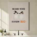 Gallery wrap stretched canvas of two cute Halloween ghosts wearing black witch hats with Wash Your Hands Boo orange typography hung frameless above a bathroom vanity with black faucet and soap dispenser