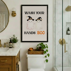 Black framed funny Halloween ghost Wash Your Hands Boo bathroom canvas art hung on a white wall between a marble vanity with gold faucet and a glass shower enclosure with wicker tray and trailing plants on toilet