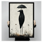 Person holding a large black framed print of a surreal gothic crow on stilted legs carrying an umbrella amid wildflowers, shown against a neutral gray background
