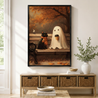 Black framed whimsical Halloween oil painting of a black cat with an orange scarf and a ghost holding a coffee cup sitting on an autumn park bench, mounted above a wicker storage console with white ceramic vases and stacked books in a warm minimal entryway