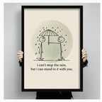 Close-up of hands holding black framed wall art print — stick figures sharing umbrella in rain with heartfelt quote on cream background