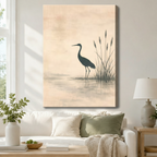 Stretched canvas wall art of a serene Japanese sumi-e style heron standing gracefully in a still misty lake beside tall slender reeds with a soft water reflection on a warm blush peach aged paper background, hung above a white sofa with sage green and cream pillows and a ceramic table lamp in a bright airy living room