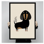 Person holding a large black framed minimalist dachshund canvas art print showing a charming flat geometric black and tan wiener dog standing beside a delicate olive branch illustration on a clean cream background against a light gray studio backdrop
