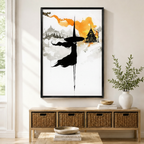 Black framed minimalist ink watercolor flying witch canvas art with a black-cloaked witch in a pointed hat soaring through gray misty mountains beside an orange-washed gothic house with dripping ink brushstrokes on a white background, displayed on a light gray wall above a wooden rattan storage console with a white vase of greenery