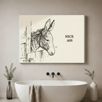 Gallery wrap stretched canvas of a pencil sketch donkey peeking through a wooden fence with Nice Ass humor quote on cream background hung frameless above a freestanding tub with black faucet and eucalyptus vases