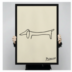 Person holding a black framed Picasso minimalist one-line dachshund sketch art print with handwritten signature on cream background against a light gray backdrop
