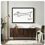Picasso-style minimalist single-line dachshund sketch black framed horizontal canvas art on white background above a dark walnut sideboard with stacked books and a succulent, beside a brass floor lamp and olive green armchair with a knit throw blanket