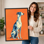 Smiling woman in cream sweater holding a black framed pop art blue and white greyhound portrait canvas art on bold orange background against a neutral indoor background with potted plants and open shelving