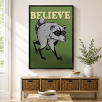 Black framed retro funny Believe raccoon canvas art with a grumpy gray raccoon with black eye mask crouching in a menacing pose and bold cream Believe lettering on a deep olive green background, displayed above a rattan storage console in a bright entryway