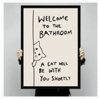 Two hands holding a large black-framed canvas print of a hand-drawn peeking cat with funny bathroom welcome quote, showing print size and frame quality