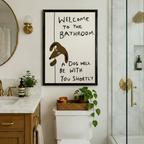 Funny dog bathroom poster framed in black hung above toilet in a warm-toned bathroom with marble countertop gold fixtures and green plants