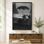Framed black and white gothic wall art print of witches relaxing on a dark beach with a black cat, displayed above a wicker sideboard in a neutral entryway